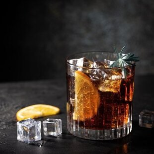 Ein Glas mit dunklem, kohlensäurehaltigem Getränk, gefüllt mit Eiswürfeln und einer Orangenscheibe, steht auf einem dunklen Untergrund. Daneben liegen zwei weitere Eiswürfel und eine Orangenscheibe. Eine dezente Garnitur, möglicherweise ein Rosmarinzweig, rundet die Präsentation ab.