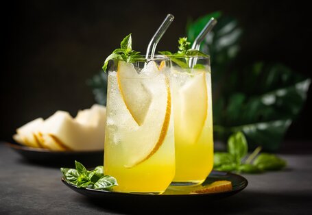 Zwei Gläser mit einem gelben, erfrischenden Getränk, garniert mit Basilikumblättern, Orangenscheiben und Melonenstücken. Die Gläser sind mit transparenten Strohhalmen versehen und stehen auf einem dunklen Teller. Im Hintergrund sind verschwommene grüne Blätter und geschnittene Melonenstücke sichtbar. Das Bild vermittelt eine sommerliche und erfrischende Atmosphäre.