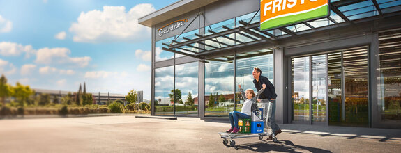 Dieses Bild zeigt den Eingang eines FRISTO Getränkemarktes, wo ein junger Vater mit seiner Tochter nach einem erfolgreichen und tollen Einkaufserlebnis auf einem Einkaufswagen den Markt verlassen.